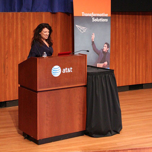 A woman standing at the podium in front of an orange and black banner.