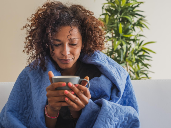 Sick woman wrapped in a blue blanket and holding a mug.