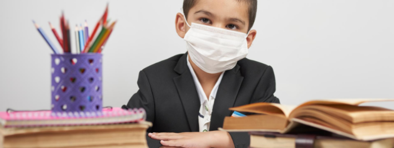 boy sitting at a desk surrounded by school books and wearing a mask