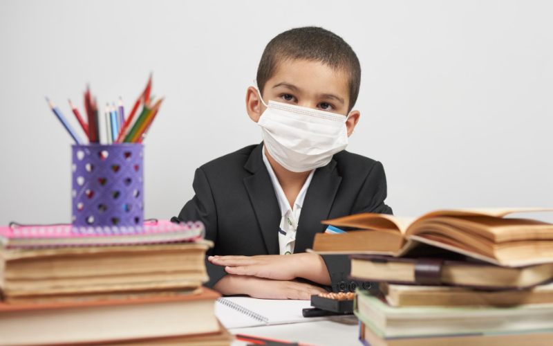 boy sitting at a desk surrounded by school books and wearing a mask
